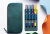 Set of colorful pens in a teal case on a white background with colorful paper pieces.